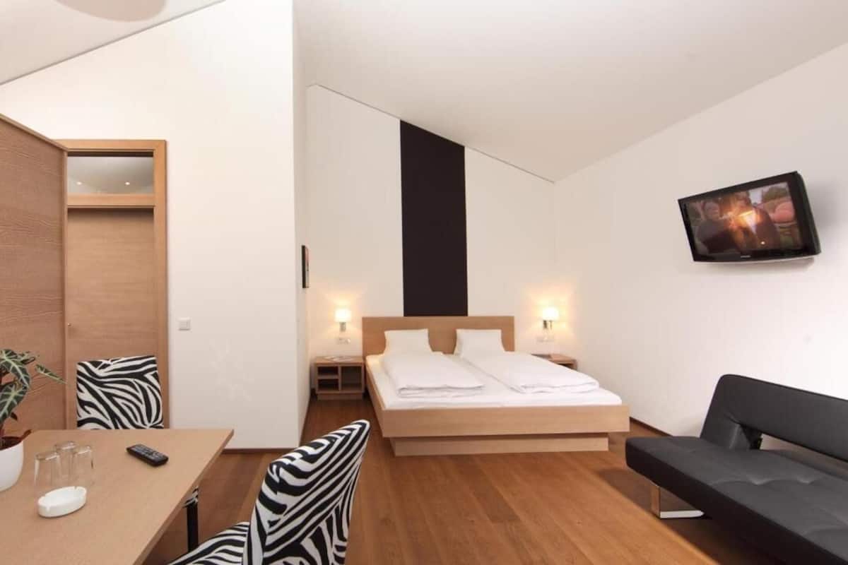 Une chambre d’hôtel moderne équipée d’un lit, d’un canapé, d’un bureau agrémenté d’une plante et d’un téléviseur fixé au mur.