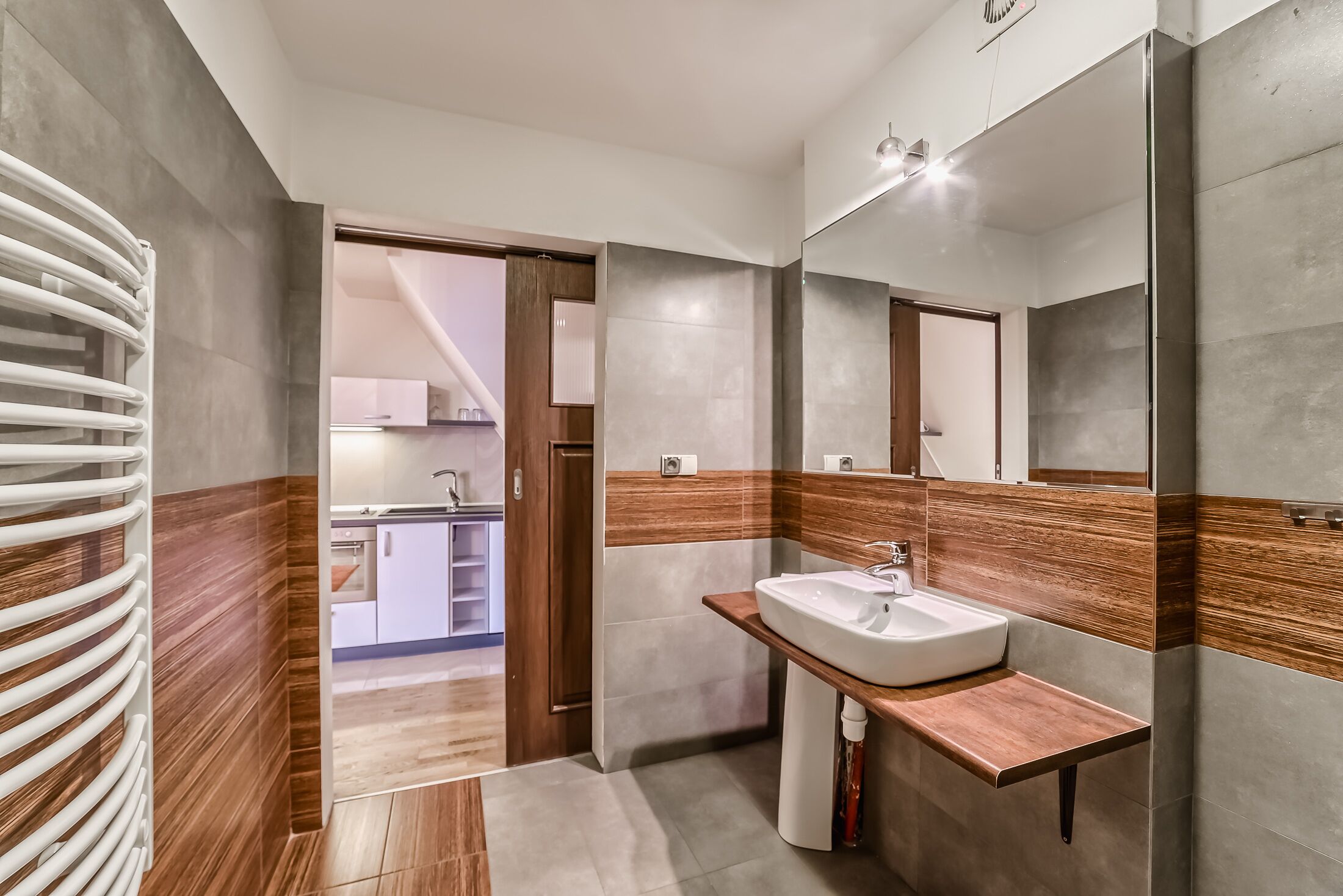A modern bathroom with a wooden vanity, a large mirror, and a view into the kitchen.