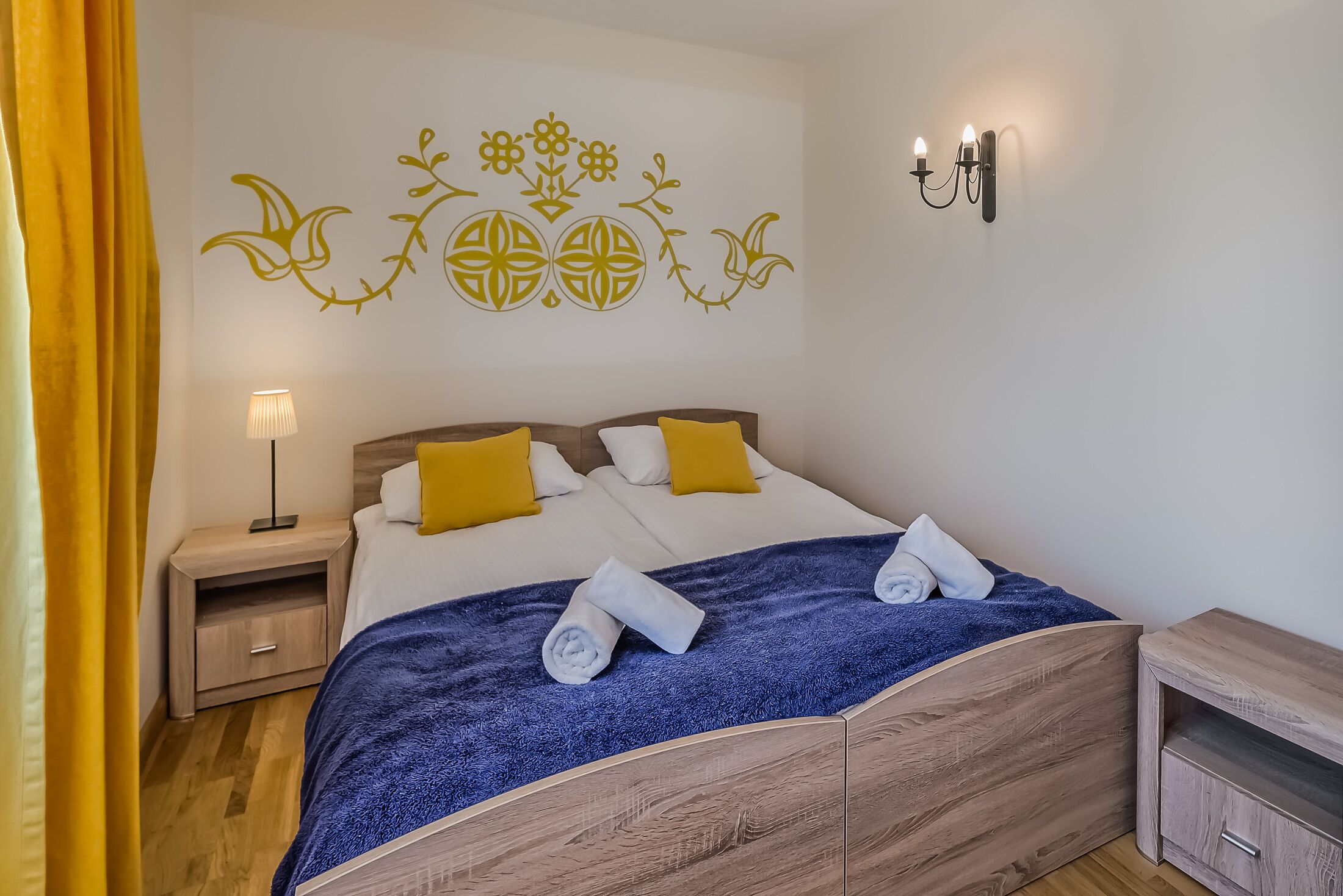 A neatly made bed with a blue blanket, two pillows, and rolled towels. There is a wall-mounted light fixture and a small bedside table with a lamp.