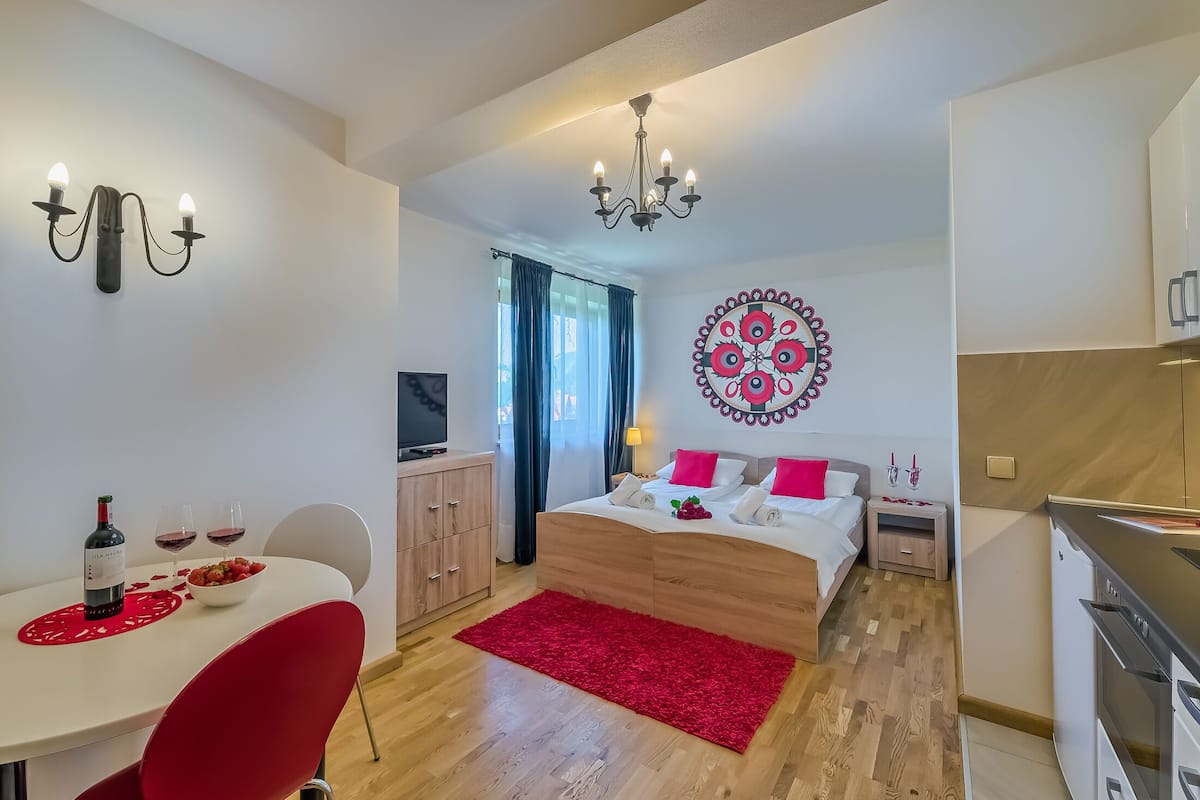 A modern bedroom with a bed, bedside table, and a red rug. There is a dining area with a table and chairs, and a kitchen area with a stove.