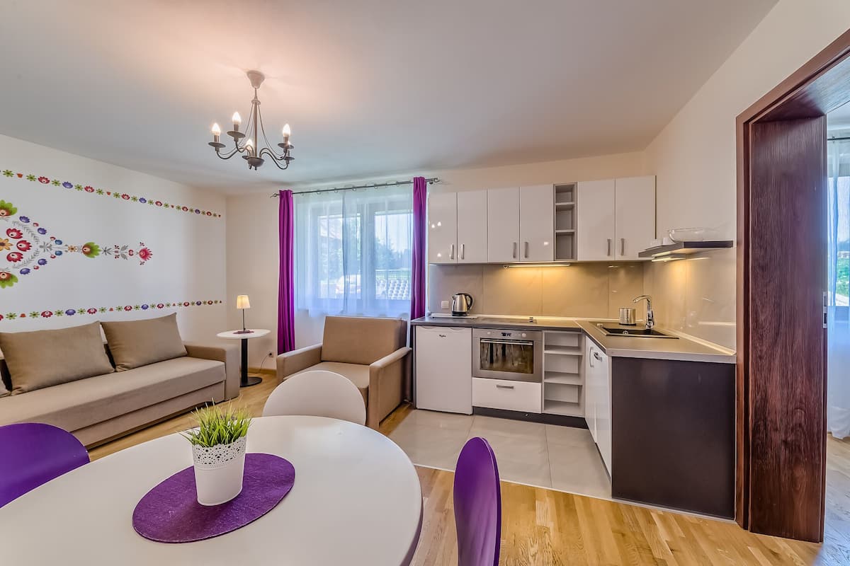 A modern kitchen with white cabinets, a built-in oven, and a sink. There is a dining area with a white table and purple chairs.