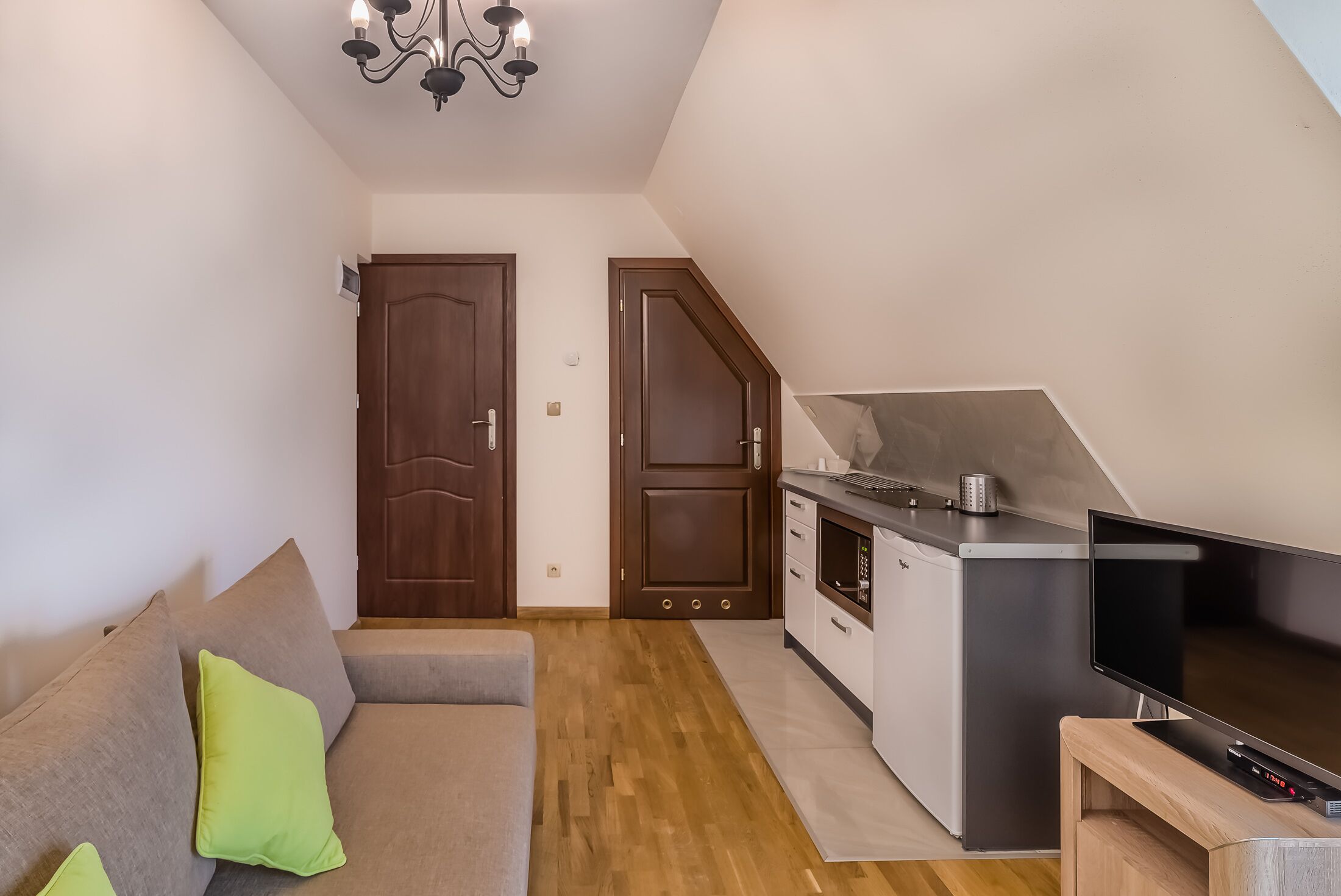 Ett kompakt bostadsutrymme med ett litet kök, en soffa med kuddar, en TV och en kristallkrona.