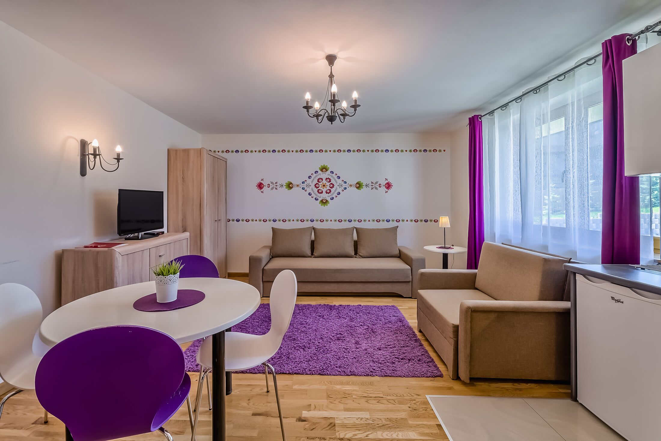 A modern living room with a sofa, a dining table with purple chairs, a TV, and a kitchen area with a refrigerator.
