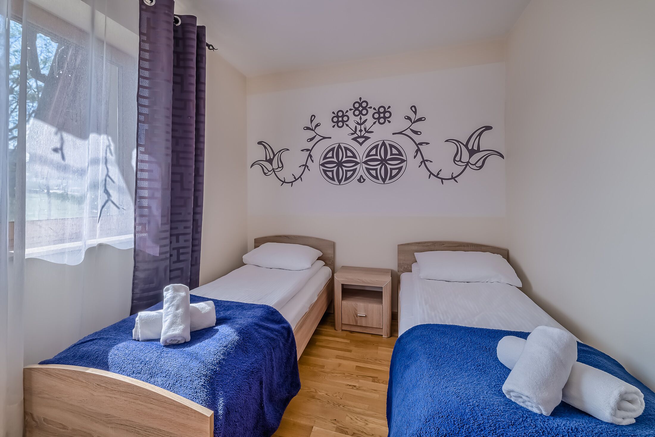 A room with two beds, each with a blue blanket and white towels, wooden headboards, and a small nightstand.
