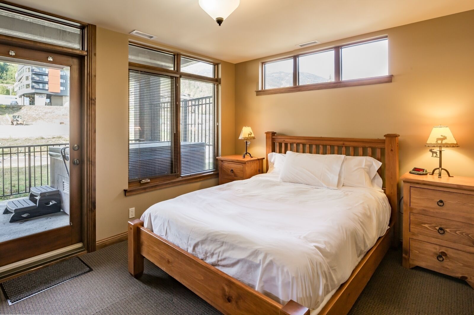 A bedroom with a wooden bed, two bedside tables with lamps, a window with blinds, and a view of a building outside.