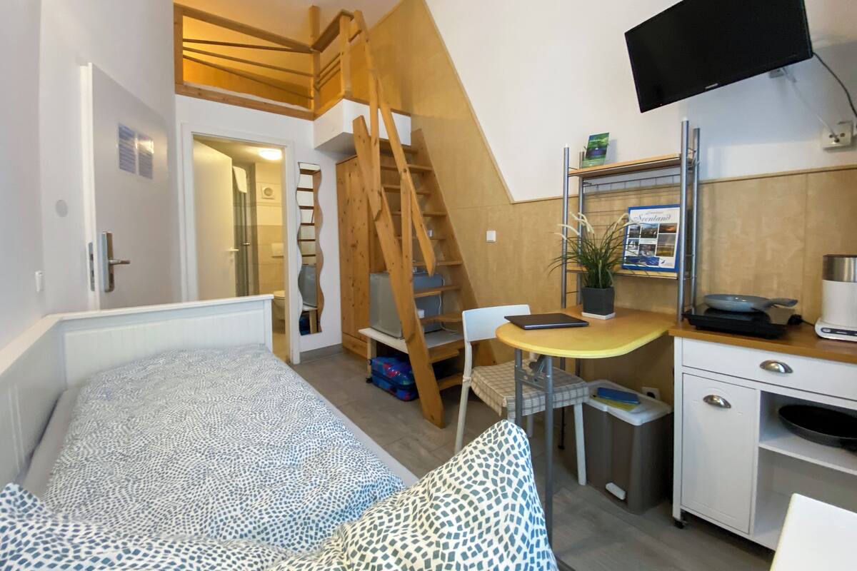 A compact hotel room with a bed, a small desk with a chair, a TV, and a kitchenette.