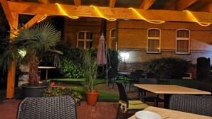 An outdoor patio with wooden beams and string lights, surrounded by potted plants and outdoor furniture. - Apartments am Schloßpark (Senftenberg)