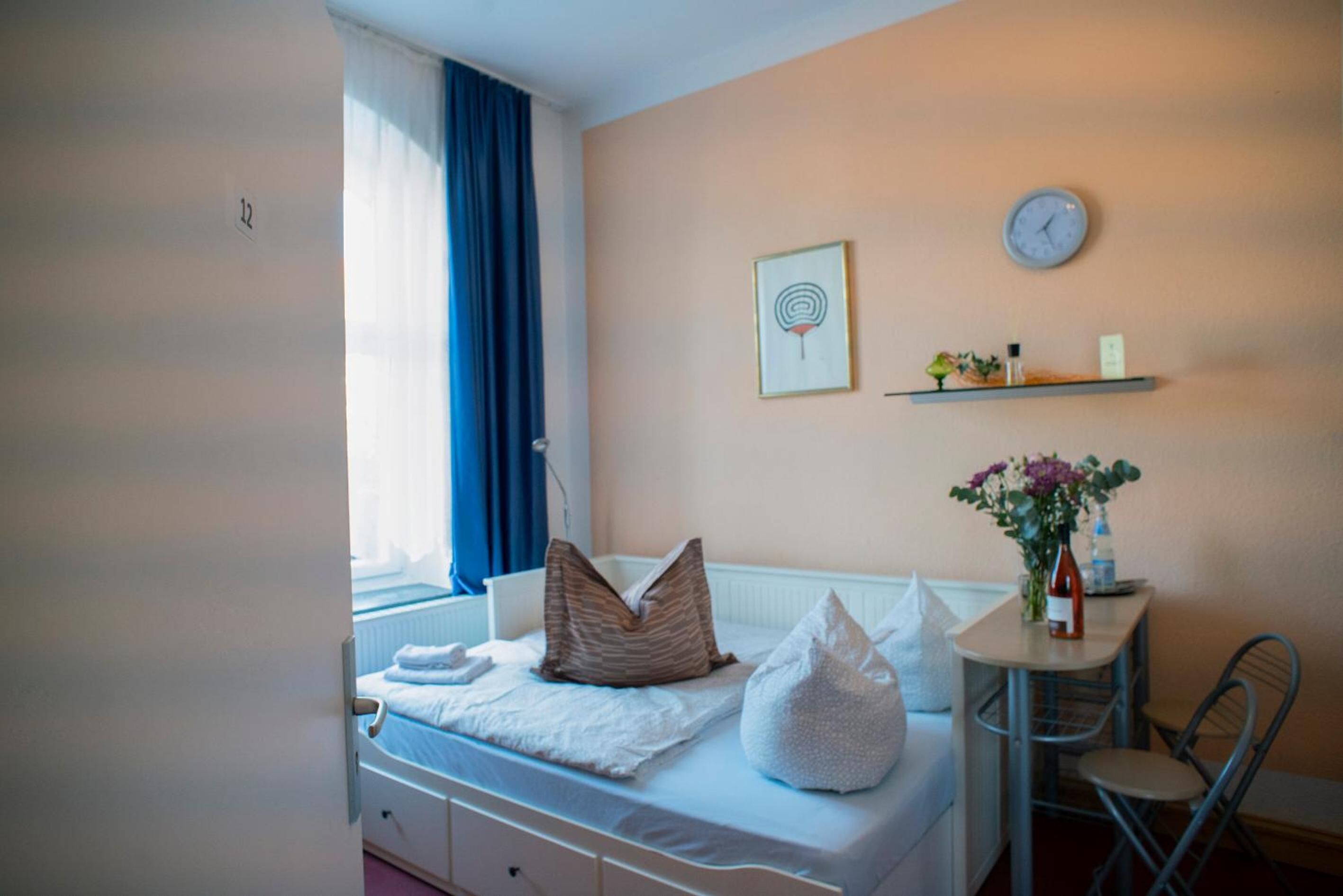 Un dormitorio con una cama, una mesita de noche con un jarrón de flores, un reloj en la pared y una ventana con cortinas.