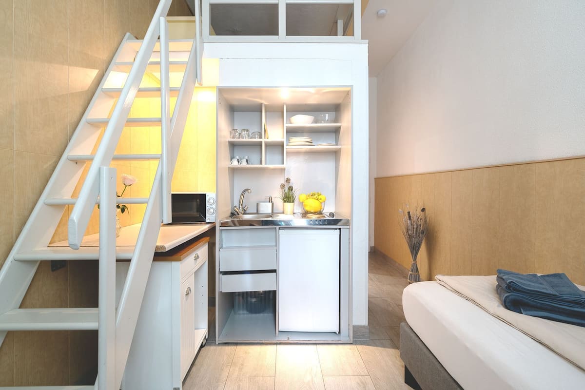 A compact kitchenette with a microwave, sink, and cabinets, a small refrigerator, and a staircase leading to an upper level.