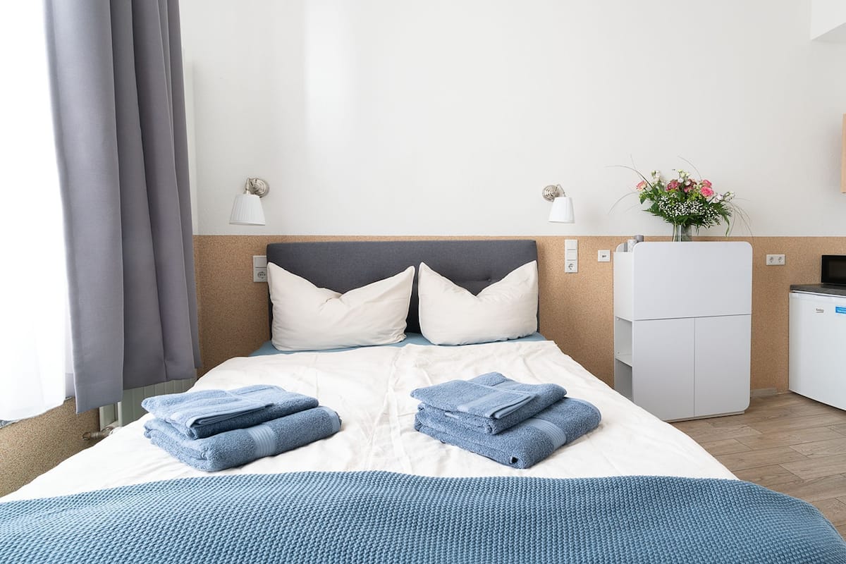 A hotel room with a bed, two pillows, a blue blanket, a white bedside table, a small refrigerator, and a flower vase.