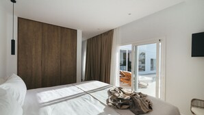 A modern hotel room with a large bed, a wooden panel wall, a sliding glass door to a balcony, and a flat-screen TV mounted on the wall.