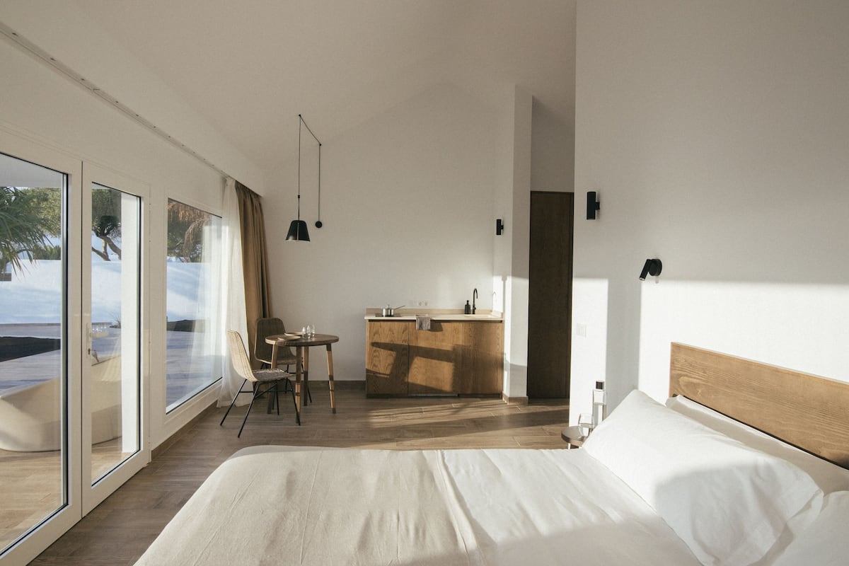 A modern hotel room with a large bed, a dining area with a table and chairs, a kitchen area, and large windows offering a view of the outdoors.