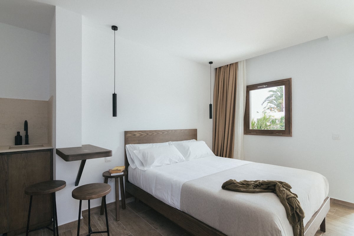 A modern hotel room with a bed, a small desk, two stools, a hanging light fixture, and a window with curtains.