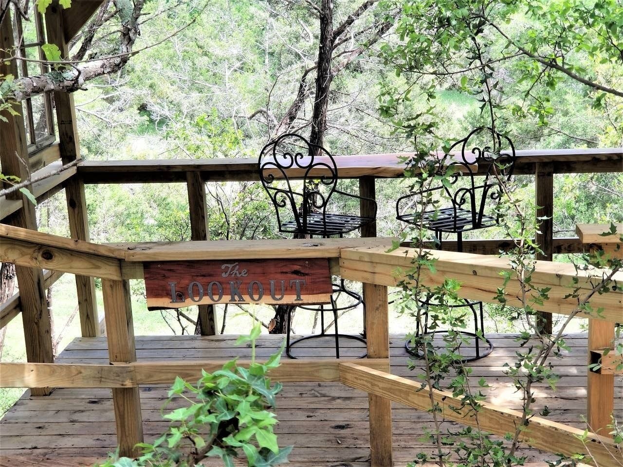 A wooden deck with a sign reading "The Lookout," surrounded by metal chairs and tables, set against a backdrop of trees.