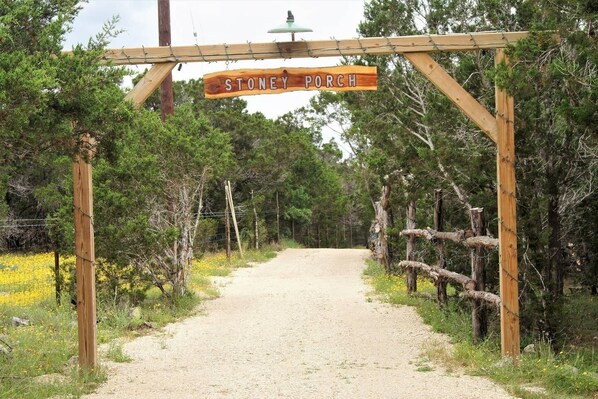 A wooden sign reading "STONEY PORCH" at the entrance of a gravel path surrounded by trees and a fence.