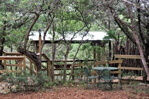 A rustic wooden gazebo with metal chairs and a table, surrounded by dense trees and a metal fence.