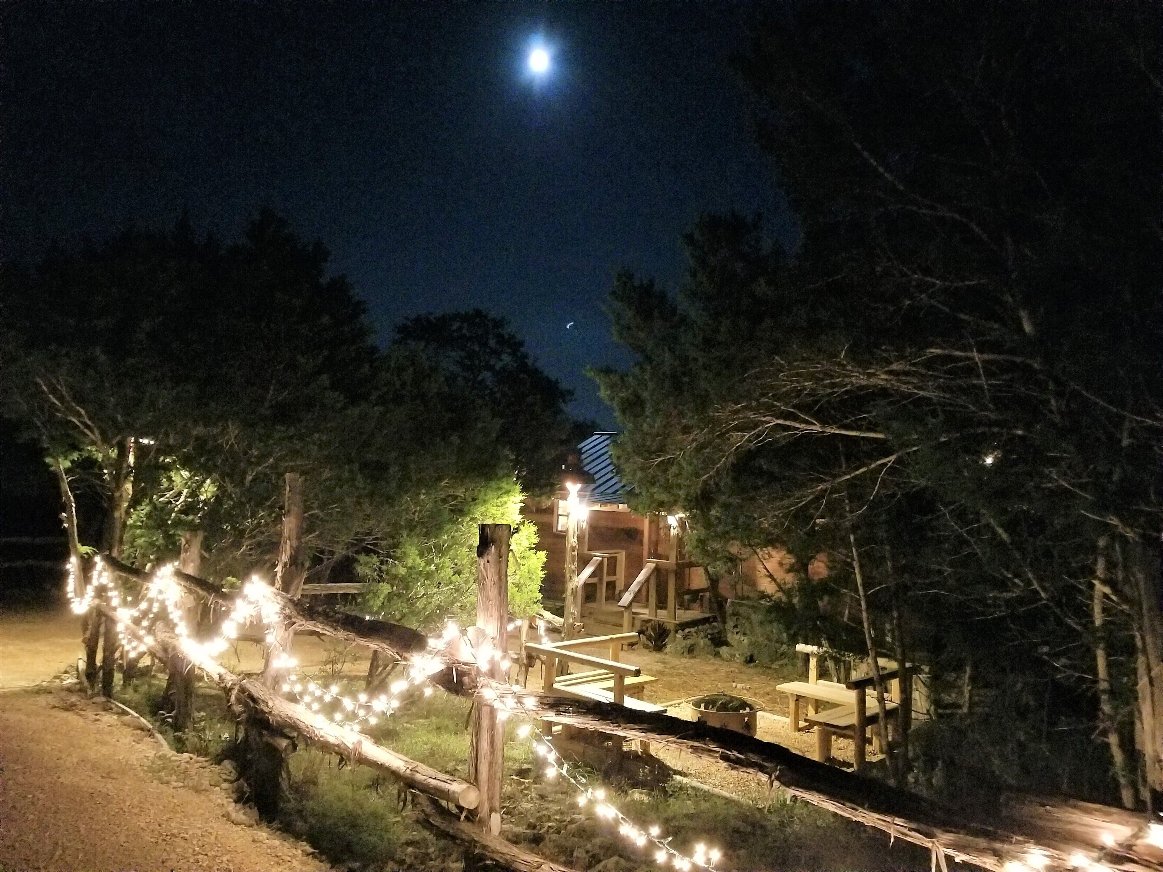 Una terraza de madera con luces colgantes, una valla de madera y un edificio con techo inclinado al fondo.