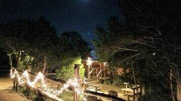 Una terraza de madera con luces colgantes, una valla de madera y un edificio con techo inclinado al fondo.