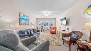 A living room with a sofa, chairs, a TV, and a ceiling fan. - TWO POOLS, FISHING PIER, PRIVATE BEACH ACCESS, COZY ACCOMMODATIONS (Gulf Shores)