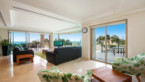 A spacious living room with a large sofa, a coffee table, and a TV. There is a sliding glass door leading to a balcony with a view of the ocean and palm trees. - Unit 13 - 3 Bedroom Ocean View (TERRIGAL)