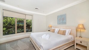 A spacious bedroom with a large window, a neatly made bed, and a nightstand with a lamp.