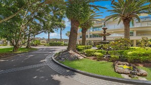 A landscaped pathway leading to a building with a fountain, surrounded by palm trees and well-maintained greenery.