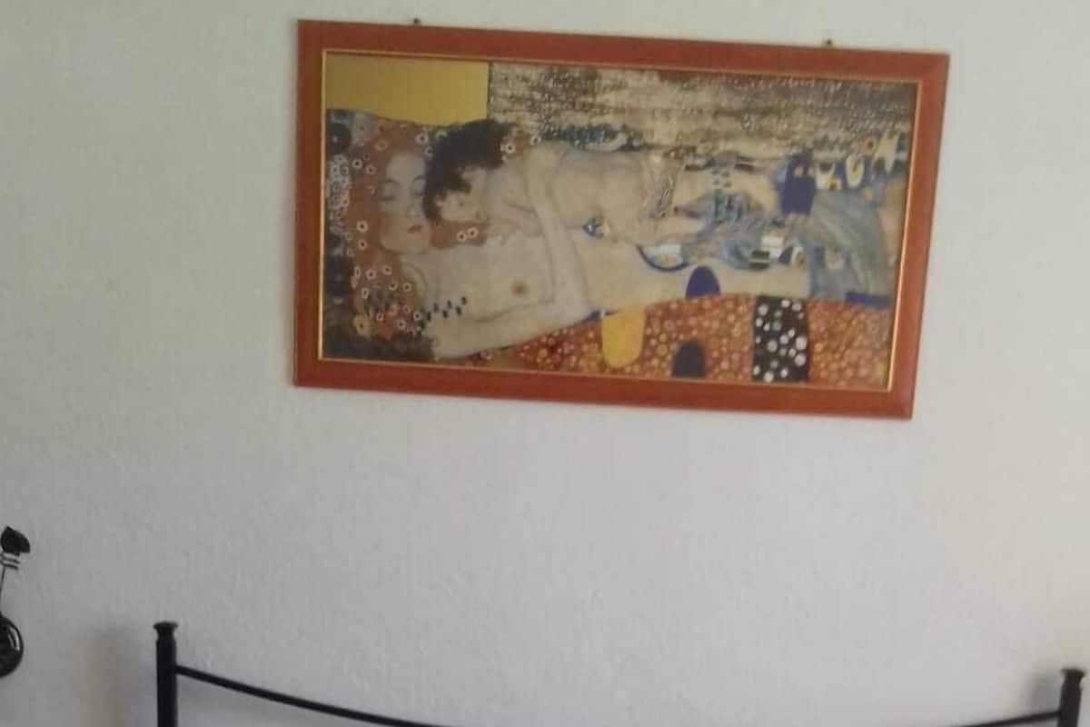 Una cama con estructura metálica y colchón gris en una habitación con pared lisa y un cuadro colgado sobre la cama.