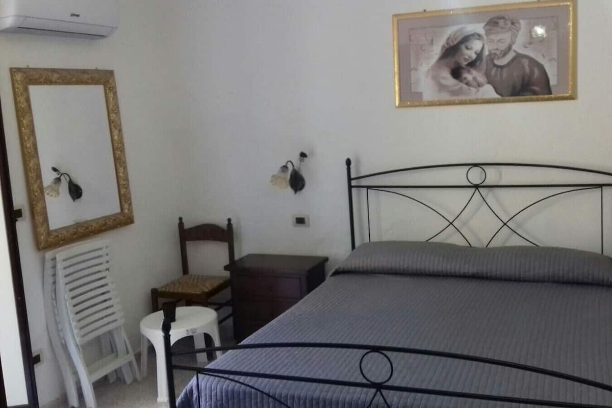 Un dormitorio con estructura de cama metálica, una mesita de noche, una silla, un escritorio pequeño y un espejo en la pared.