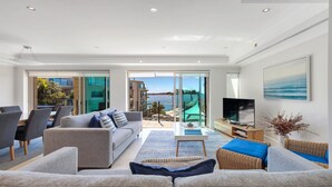 A modern living room with a sofa, a television, and a view of the sea through large glass doors. - Unit 25  - 3 Bed Ocean View (TERRIGAL)