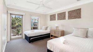 A bedroom with two beds, a ceiling fan, a nightstand, and a view of a balcony with palm trees. - Unit 26 - 3 Bedroom Premier Ocean View (TERRIGAL)