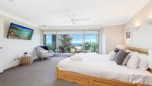 Un dormitorio con una cama grande, ventilador de techo, televisión y vista al océano a través de puertas correderas amplias.