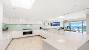 Una cocina moderna con armarios blancos, horno empotrado y una isla grande. Hay un comedor con vista al océano.