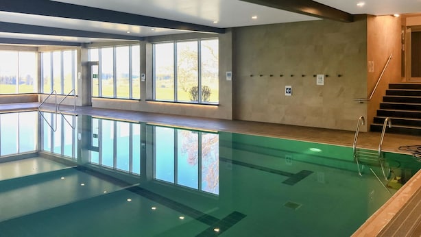 Ein Schwimmbecken mit klaren Glaswänden, einer Treppe auf der rechten Seite und großen Fenstern mit Blick ins Freie.
