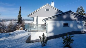 Ein modernes Haus mit einem großen Glasbalkon, umgeben von verschneiter Landschaft und Bäumen.