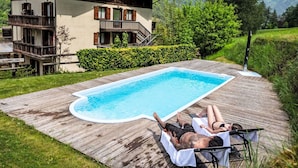 Eine Holzterrasse mit Schwimmbad, ein zweigeschossiges Chalet und eine Bergkulisse.