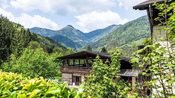 Eine Holzhütte mit Balkon, umgeben von üppiger Vegetation und Bergen im Hintergrund.