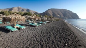 A beach with rows of sun loungers and thatched umbrellas, a clear sky, and mountains in the background.