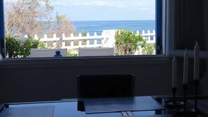 A room with a view of the ocean through a window, a desk with a chair, and a white fence in the background.