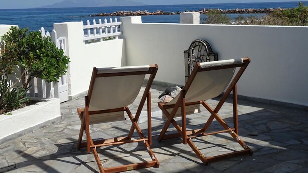 Two wooden lounge chairs with canvas seats on a stone-tiled patio overlooking the sea.