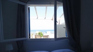A bedroom with a bed, a nightstand, a window with curtains, and a view of the beach.