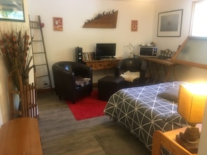 Un dormitorio con una cama, dos sillas, un escritorio con una computadora, un televisor y una alfombra roja.
