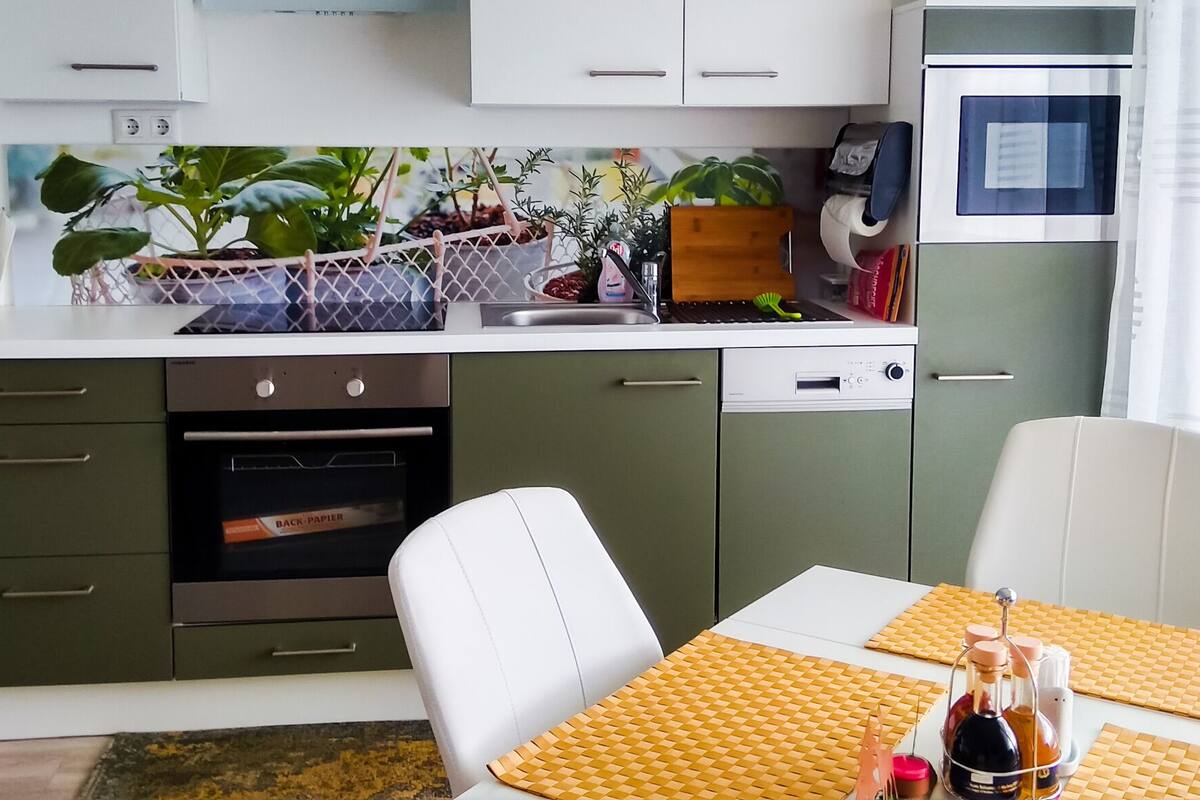 Une cuisine moderne avec des meubles verts, un plan de travail blanc et un coin repas comprenant une chaise blanche et une table.