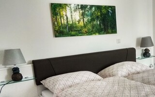 Un dormitorio con una cama, dos mesitas de noche con lámparas, un cuadro colgado en la pared que representa un bosque y una pared blanca.