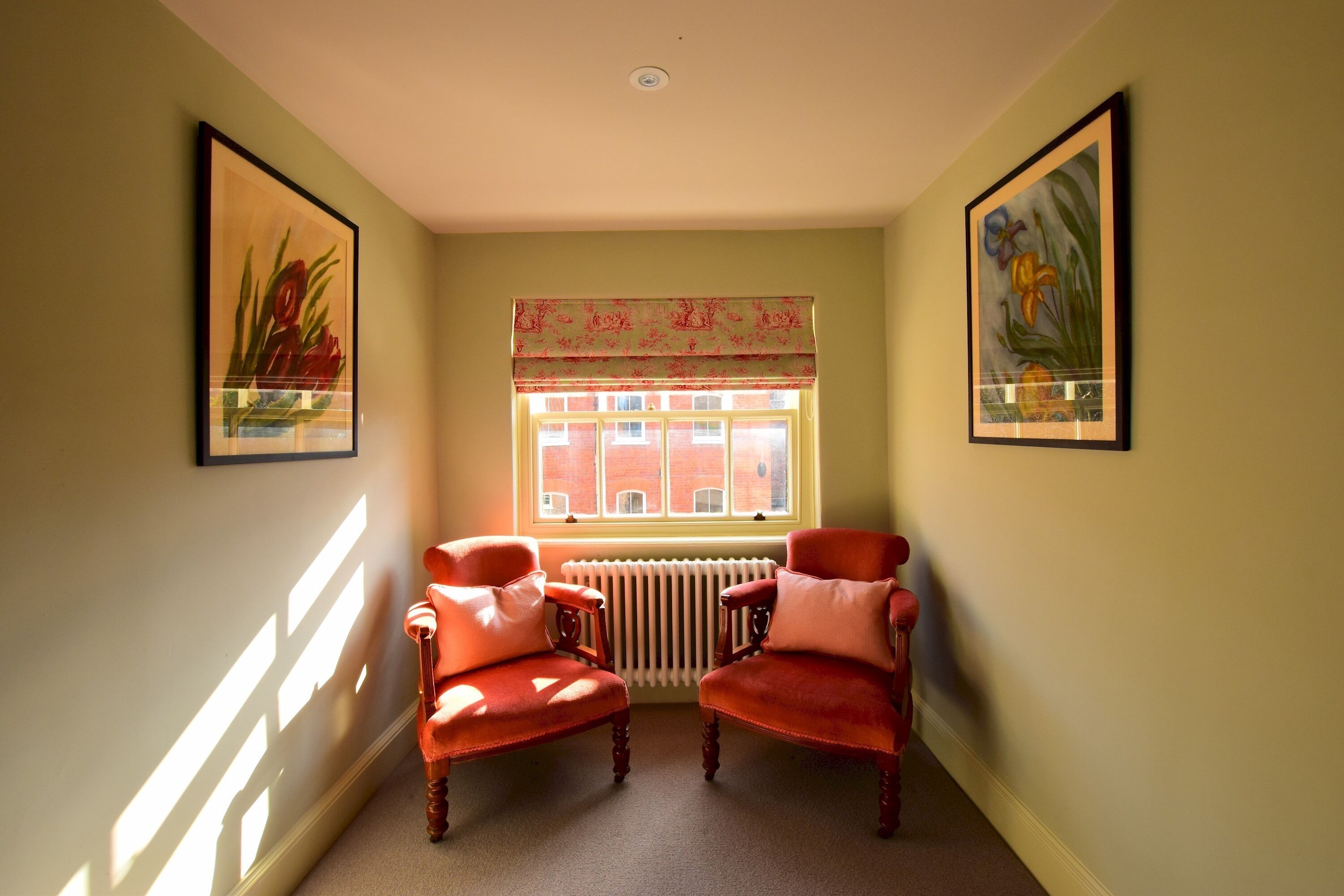 Una habitación con dos sillones rojos, un radiador, una ventana con un adornado de cortina estampado, y dos cuadros con motivos florales en las paredes.