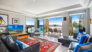 A spacious living room with a large TV, comfortable seating, and a view of the sea through large windows. - Unit 49  - 3 Bed Ocean View (TERRIGAL)