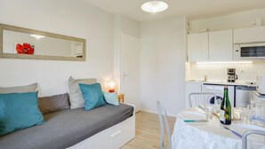 A modern, well-lit room with a sofa, a dining table set for two, and a kitchen area. - Nice apartment for 4 guests with WIFI, TV, balcony and pets allowed (Cabourg)