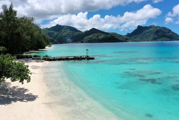 A beach with clear turquoise water, a wooden pier, and lush green hills in the background.