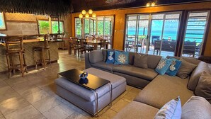 Ein großzügiger Wohnbereich mit einer großen Couch, einem Couchtisch und einer Bar mit Hockern. Große Fenster bieten einen Blick ins Freie.