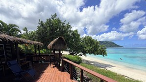 Eine Holzterrasse mit Liegestühlen und einer strohgedeckten Struktur mit Blick auf einen Strand, der klares türkisblaues Wasser und eine ferne Bergkette zeigt.
