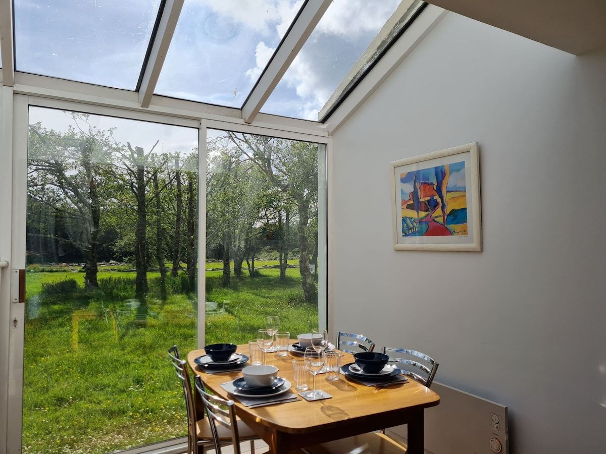 Een eetruimte met een tafel voor vier personen, een schilderij aan de muur en uitzicht op een groen landschap door een groot raam.